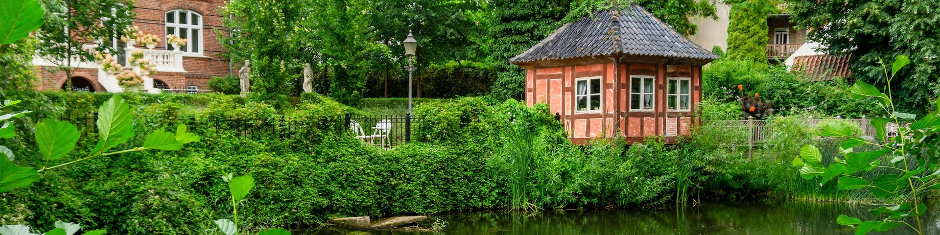 A serene pond with a small red-roofed structure surrounded by lush greenery and a brick house in the background.