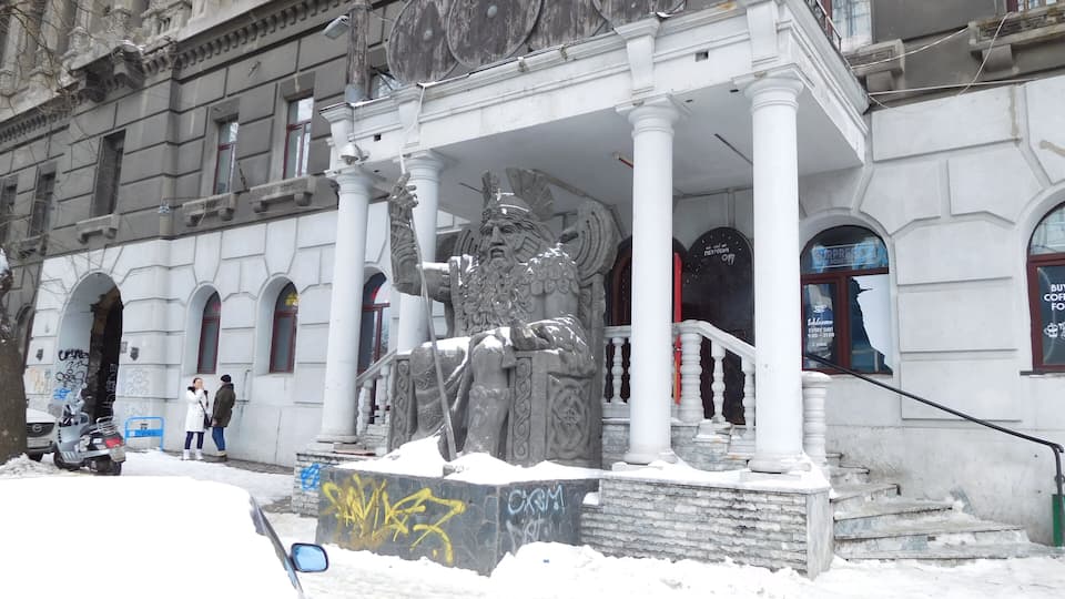 A statue in front of a building with columns and graffiti on the wall.