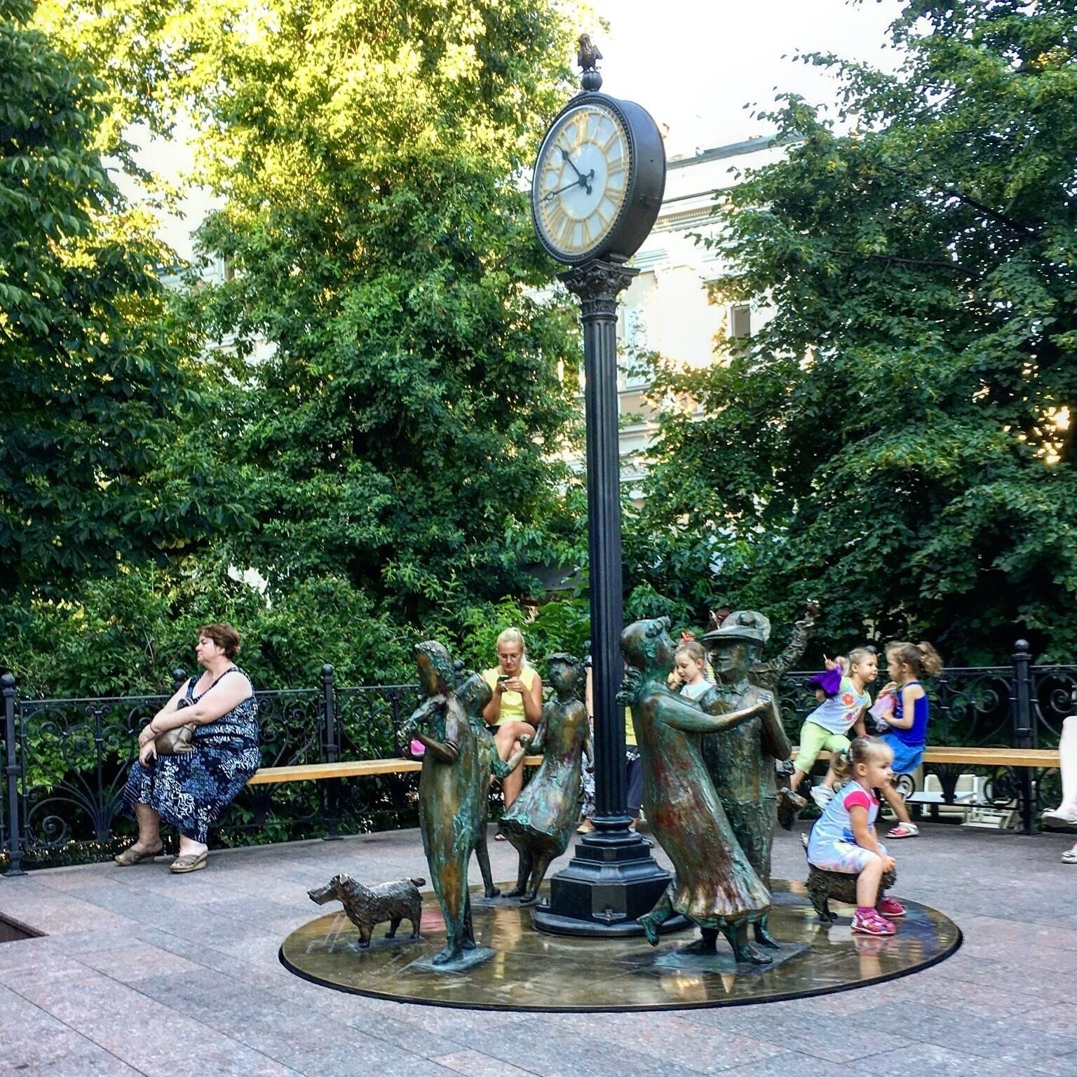 Um parque com relógio, estátuas de bronze e pessoas sentadas em bancos.