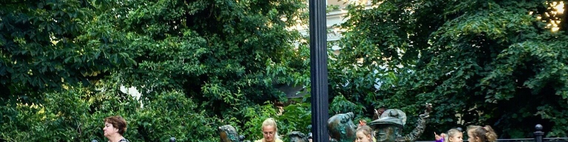 Um parque com relógio, estátuas de bronze e pessoas sentadas em bancos.