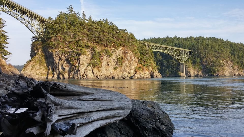 A bridge over a river with a forested cliff and a large fallen tree trunk in the foreground.