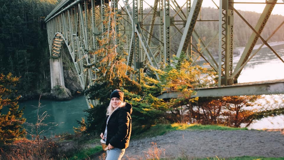 A person standing in front of a large steel bridge with a river below.