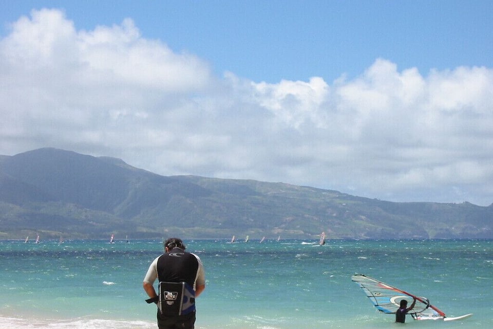 A beach with clear turquoise water, a person walking, and windsurfing equipment on the sand.