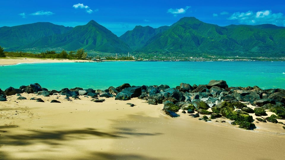 A beach with turquoise water, rocky shoreline, and green mountains in the background.