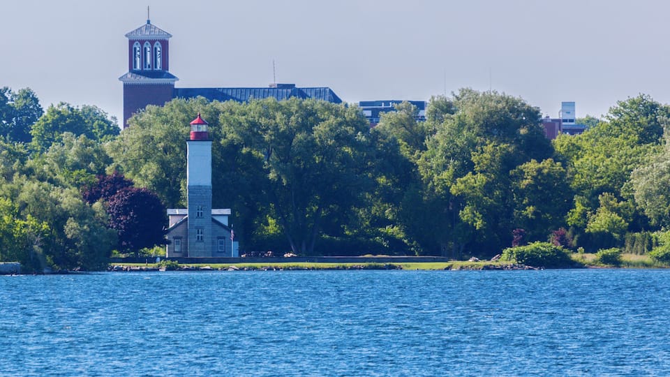 A lighthouse on a small island, surrounded by dense trees and a large building in the background.
