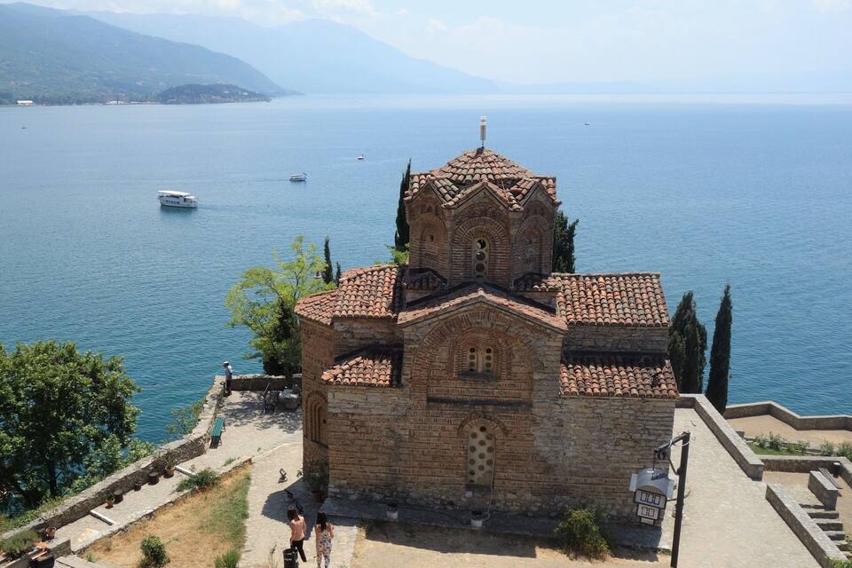 A stone church with a tiled roof by a lake, surrounded by trees and a clear sky.