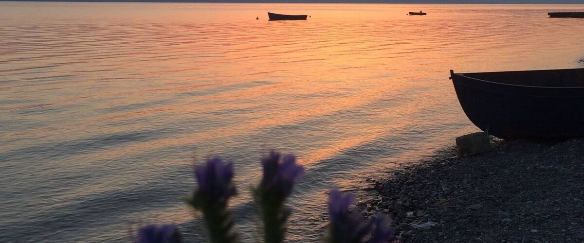A sunset over a calm lake with boats and a rocky shore.