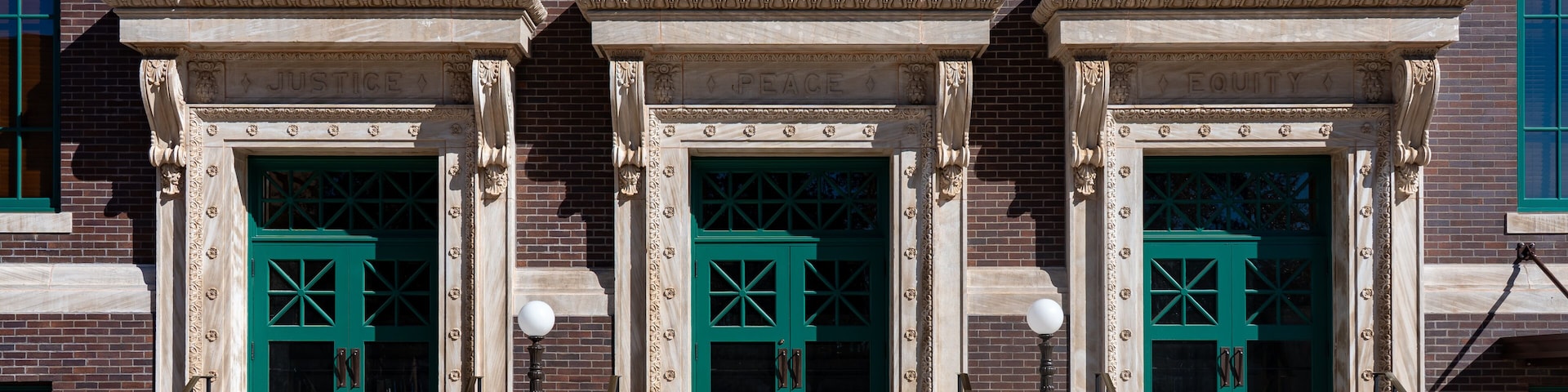 Un bâtiment doté de trois portes vertes, chacune encadrée par une colonne de style classique et surmontée d’un fronton orné d’inscriptions.