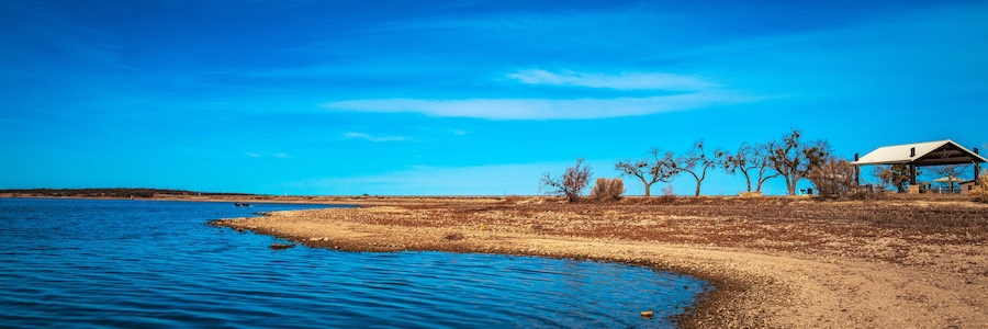 A serene lakeside scene with a sandy shore, a small building, and leafless trees under a clear blue sky.