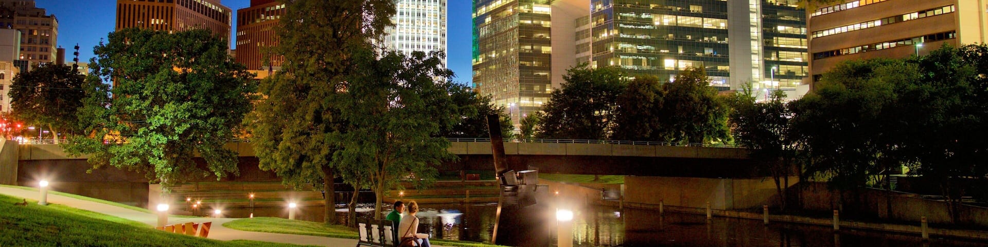 A cityscape at night with illuminated buildings, a river, and a park with people walking.