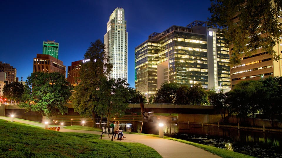 A cityscape at night with illuminated buildings, a river, and a park with people walking.