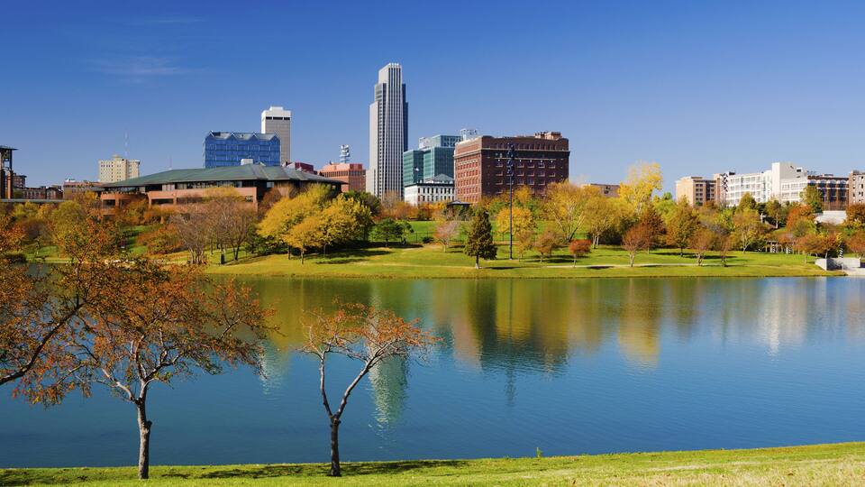 A lake with trees in autumn colors, surrounded by a park and city buildings in the background.