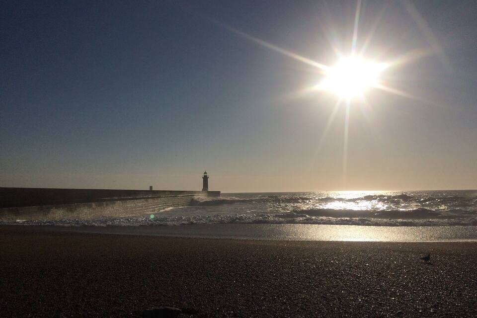 A beach with waves, a lighthouse, and the sun shining.