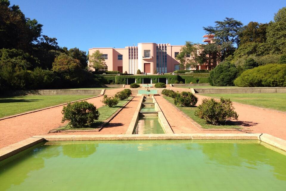 A formal garden with a central water feature, geometric pathways, and a large building in the background.