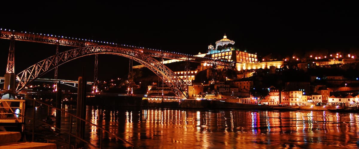 A nighttime view of a bridge over water, with illuminated buildings and a hill in the background.
