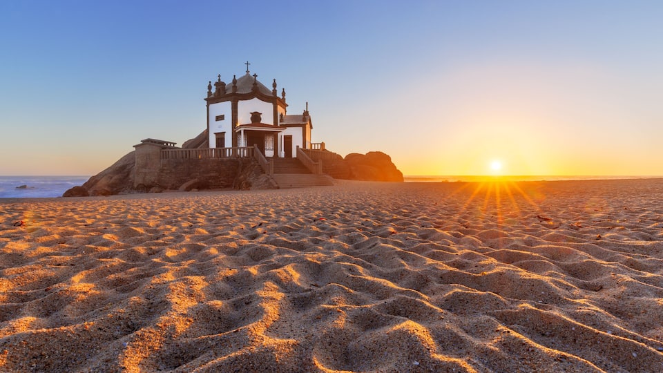A white church on a rocky outcrop with the ocean in the background and the sun setting.