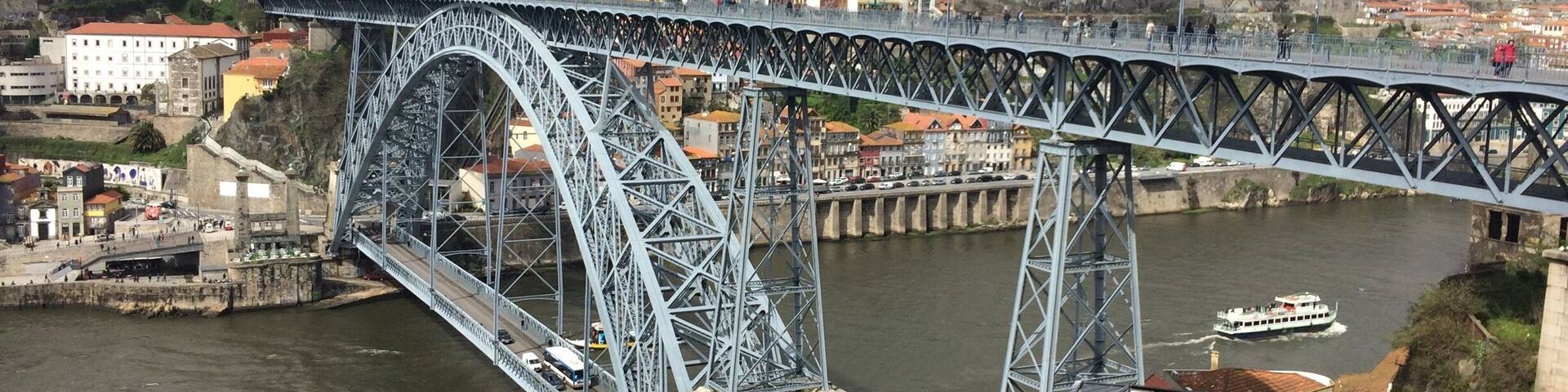 A large steel bridge spanning a river, with buildings and a boat in the background.