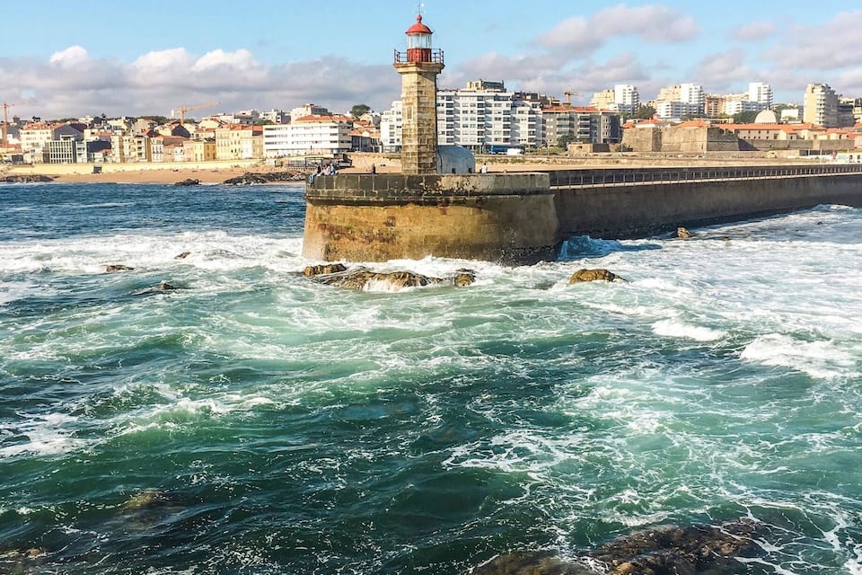 A coastal cityscape with a prominent lighthouse on a breakwater, waves crashing against the shore, and a backdrop of buildings.