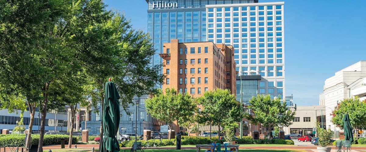A park with green trees, a statue, and outdoor seating. In the background, there are modern buildings including a Hilton hotel.