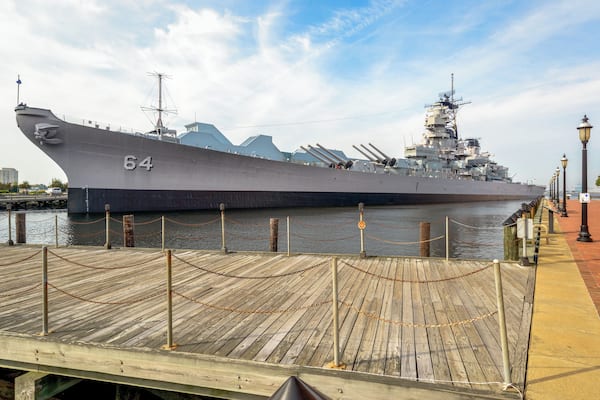 A large naval ship docked at a pier with a wooden walkway and a clear sky.