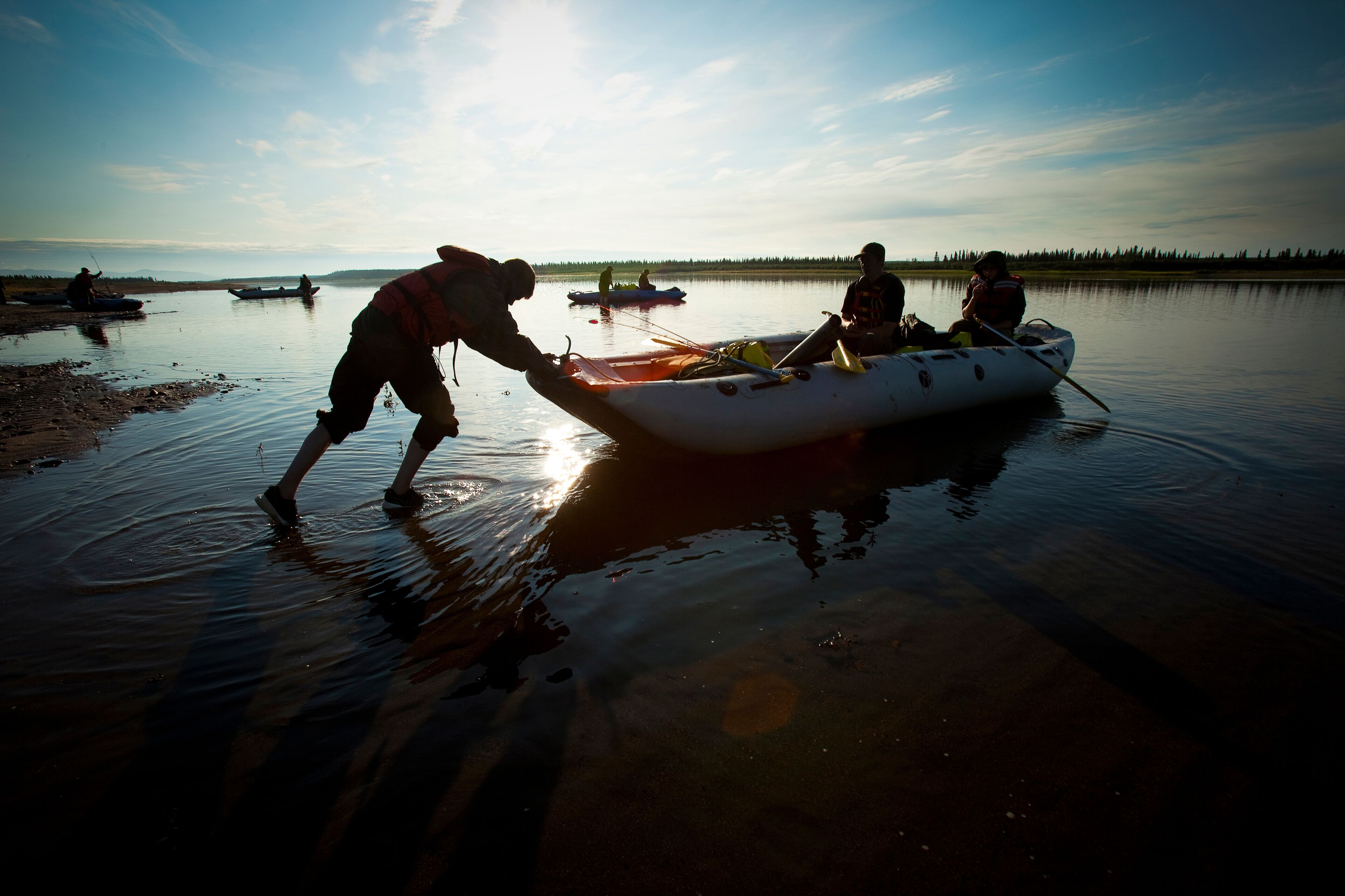 Silhouette of a boy pushing a raft with his friends away from the river bank.