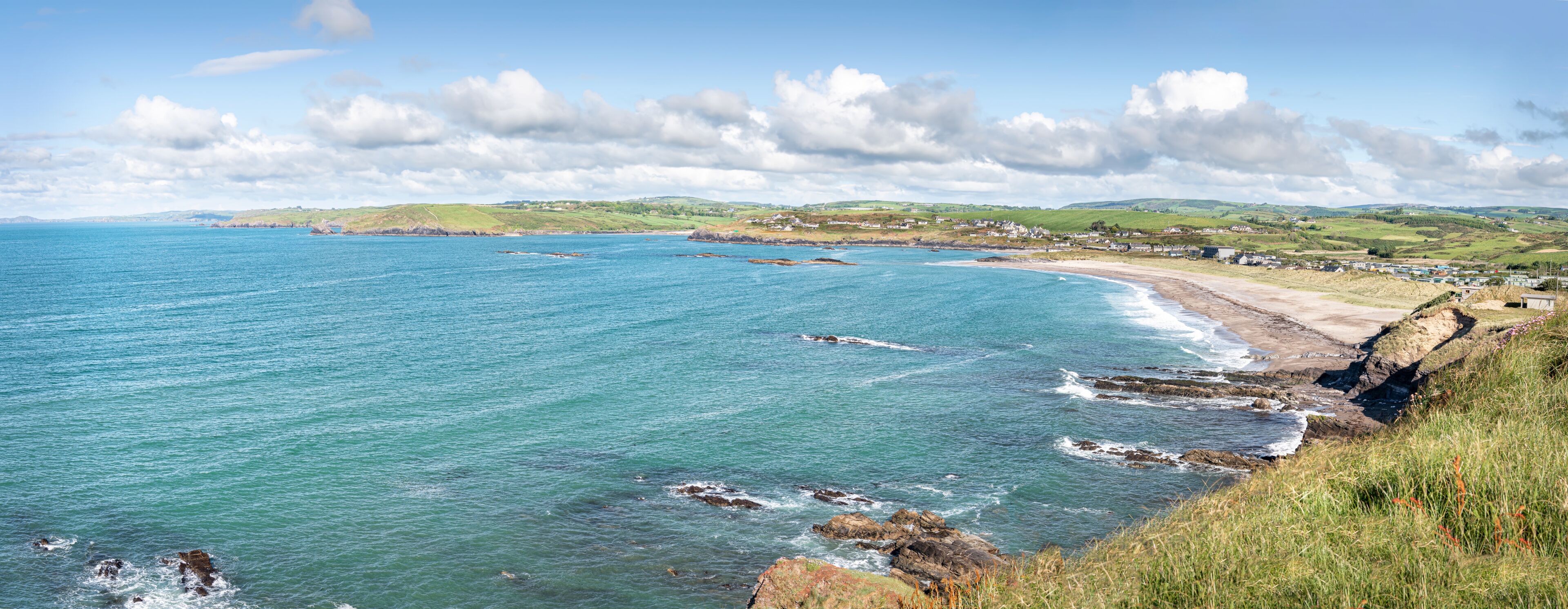 A coastal landscape with a clear blue sea, rocky shoreline, and a distant village.