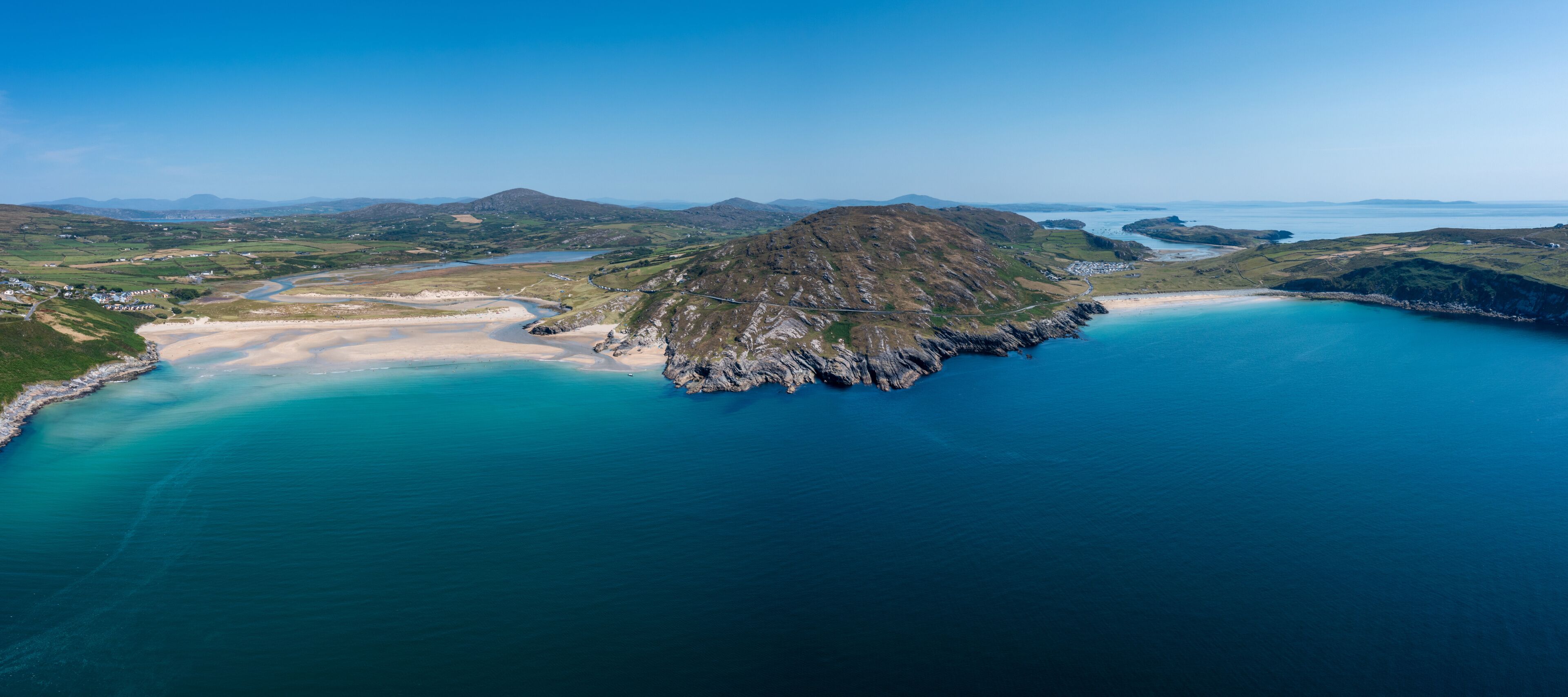 A coastal landscape with a prominent rocky island surrounded by turquoise waters, a sandy beach, and distant hills.