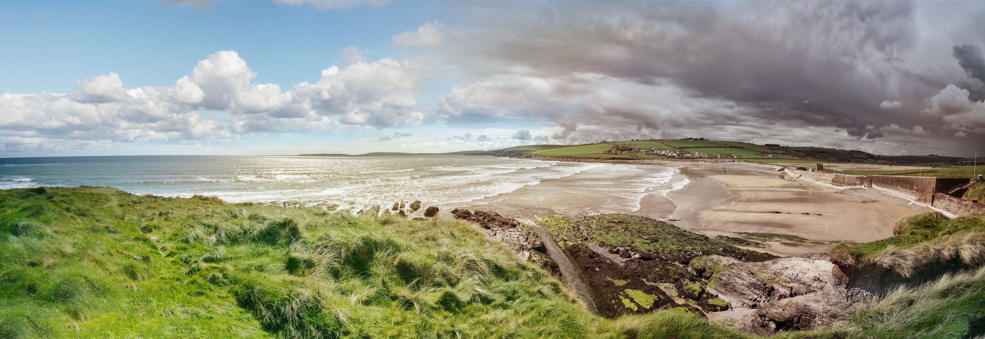 A coastal landscape with grassy cliffs, a sandy beach, and a distant village.