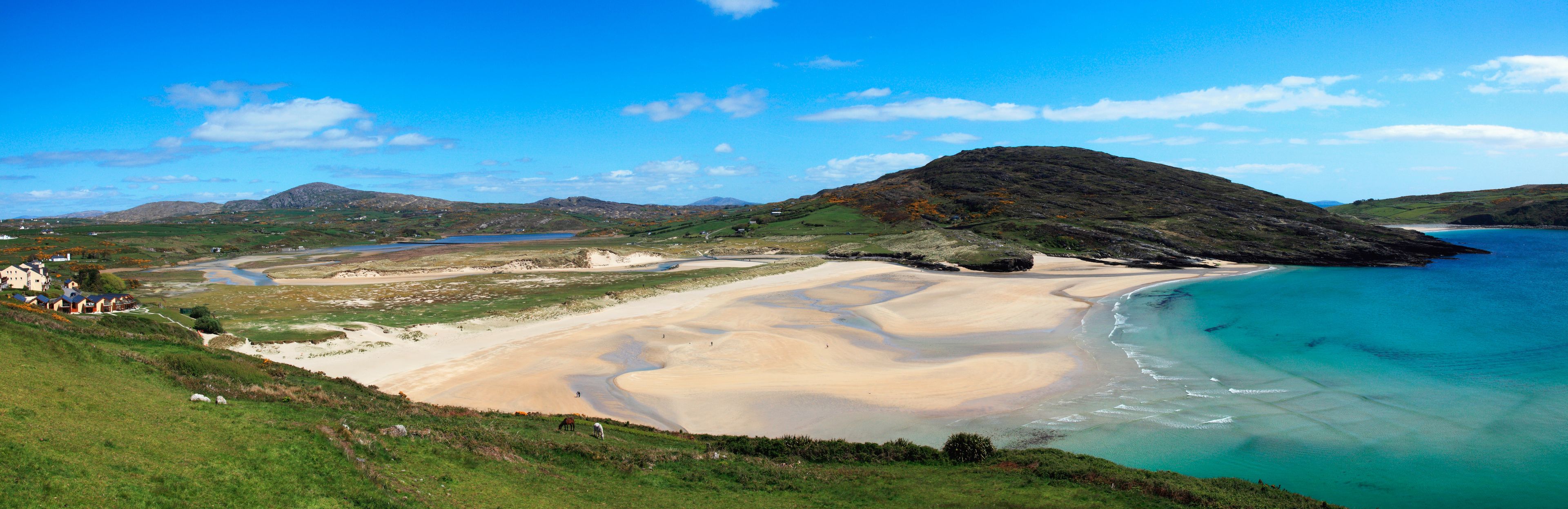 A coastal landscape with a sandy beach, a small hill, and a clear blue sky.
