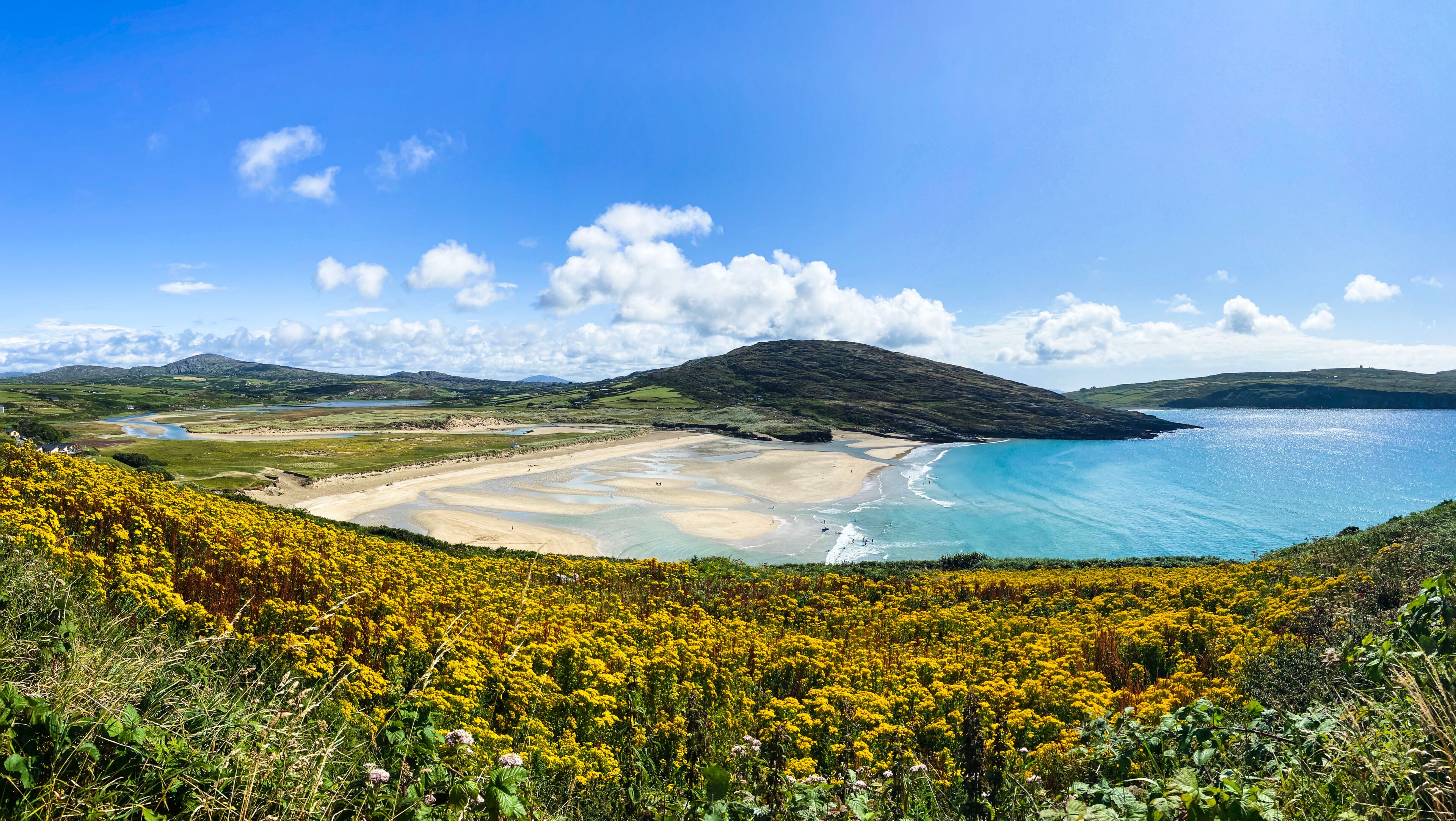 A coastal landscape with a sandy beach, clear blue waters, and a hill with vegetation.