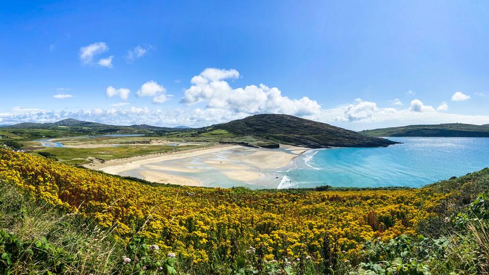 A coastal landscape with a sandy beach, clear blue waters, and a hill with vegetation.
