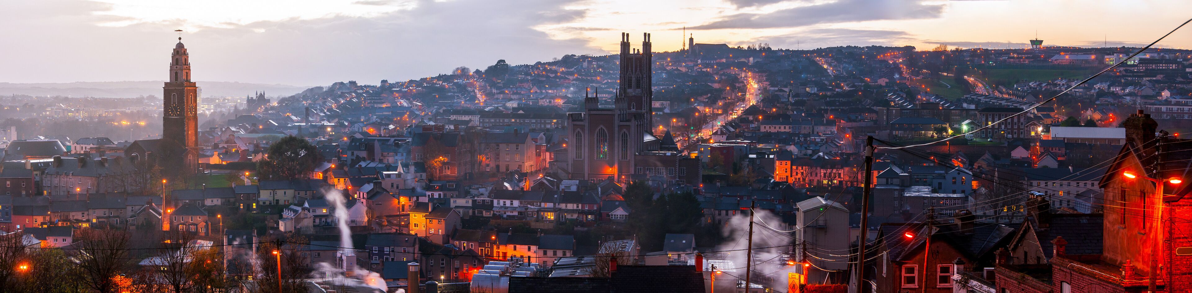A cityscape at dusk with illuminated buildings, a prominent clock tower, and a hilltop church.