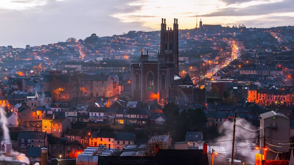 A cityscape at dusk with illuminated buildings, a prominent clock tower, and a hilltop church.