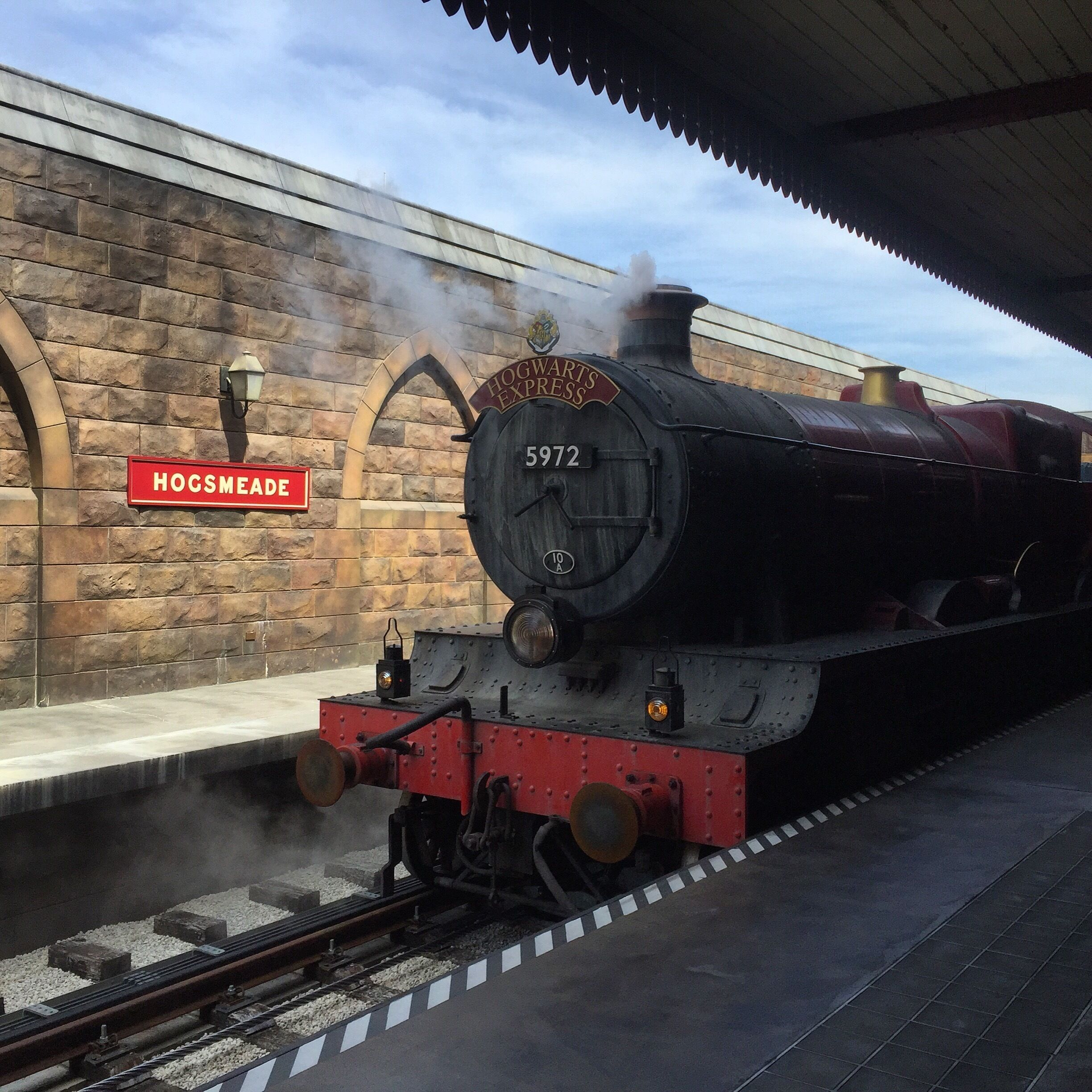 Eine Dampflokomotive an einem Bahnhof mit einer Steinmauer und einem Schild, auf dem "Hogsmeade" steht.