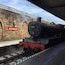 A steam locomotive at a station with a stone wall and a sign reading "Hogsmeade".