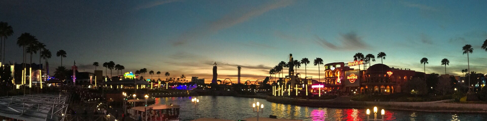 Una zona frente al mar con barcos amarrados, palmeras y edificios iluminados al anochecer.