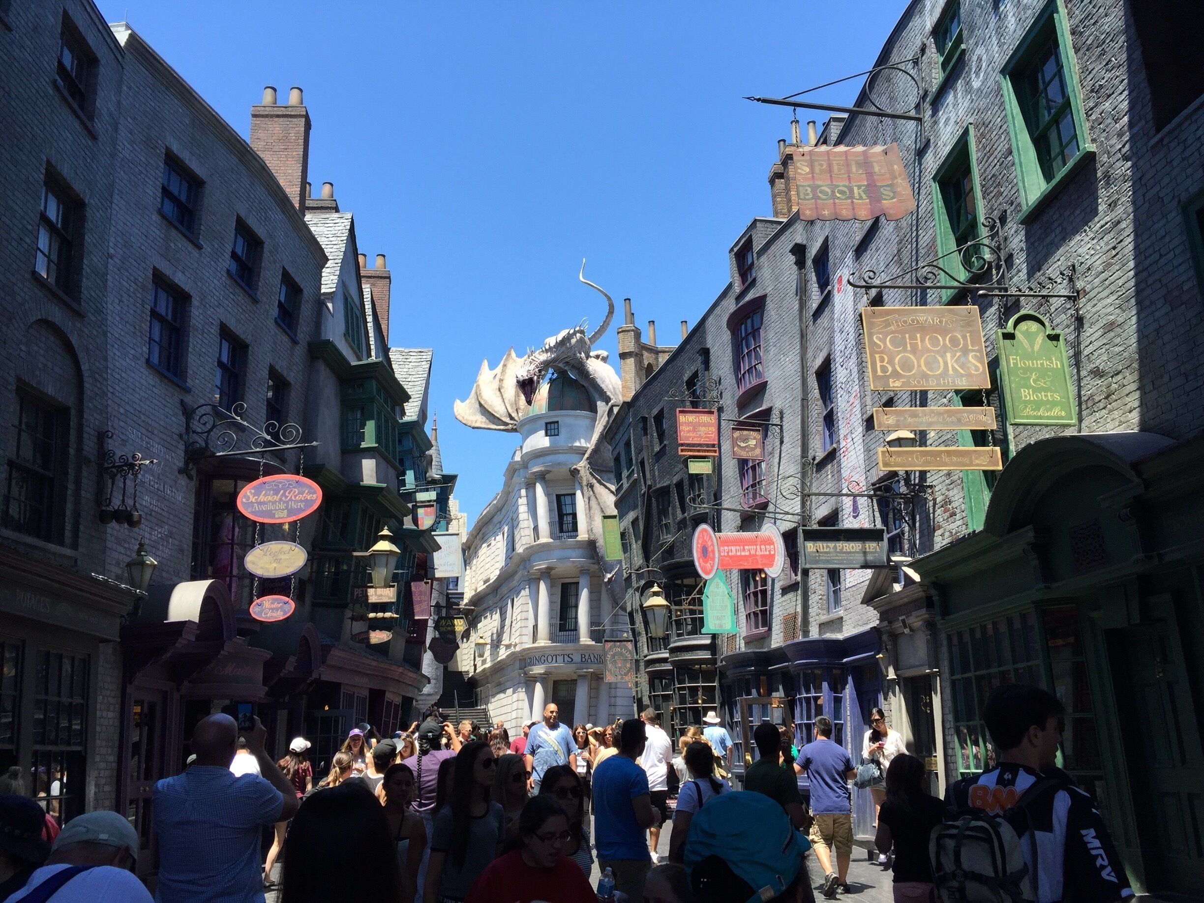 Una calle animada flanqueada por edificios de ladrillo, algunos con contraventanas verdes y letreros de tiendas como 'Spell Books' y 'Daily Prophet'.