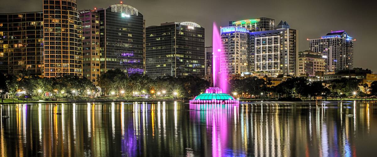 A cityscape at night with illuminated buildings and a fountain reflecting in the water.