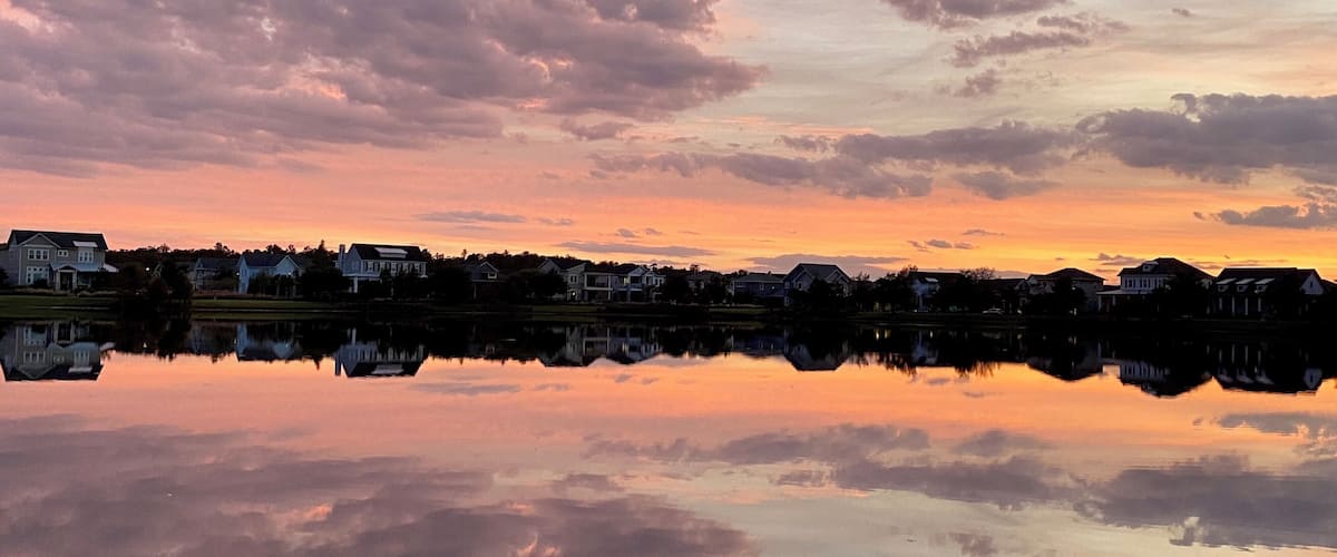 A sunset over a residential area with houses reflected in a calm lake.