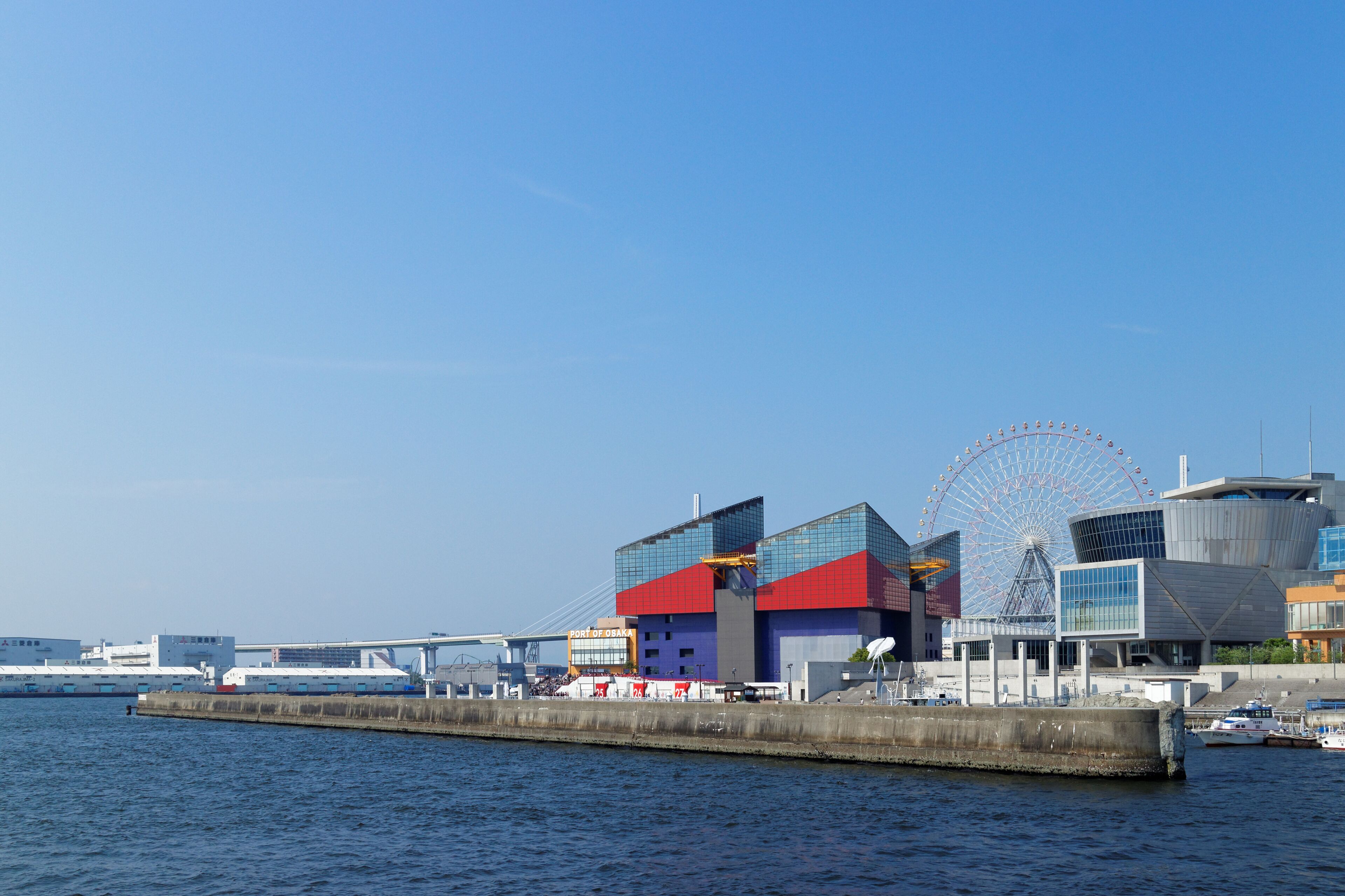 Un muelle con un edificio moderno, una noria y un puente al fondo.