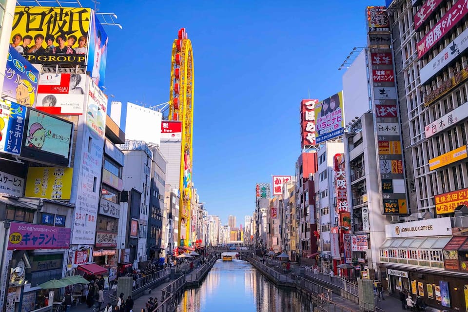 A busy urban street with a canal in the center, surrounded by tall buildings with various signs and advertisements.