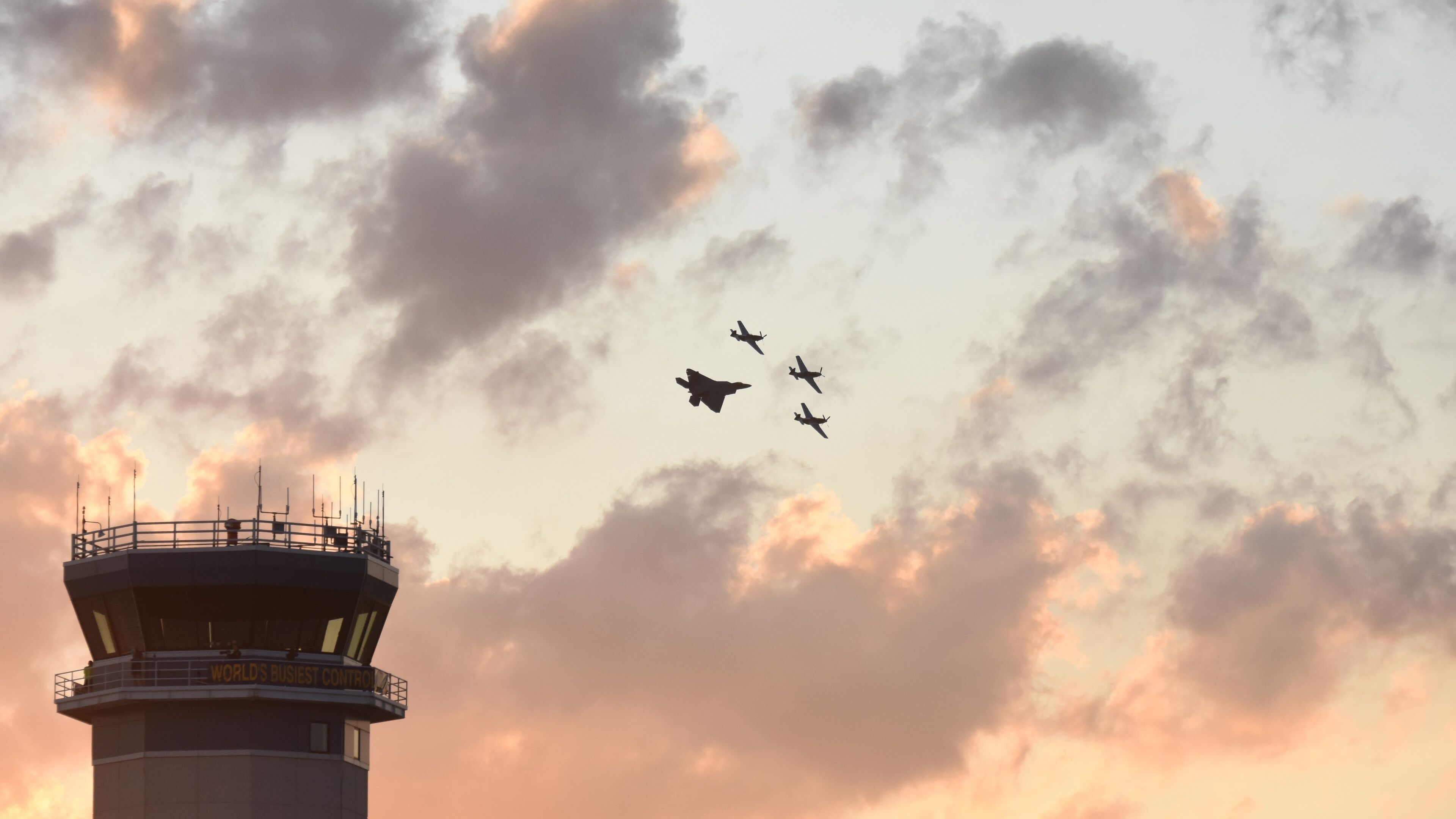 A control tower with the sign "WORLD'S Busiest Control Tower" and four airplanes flying in formation in the sky.