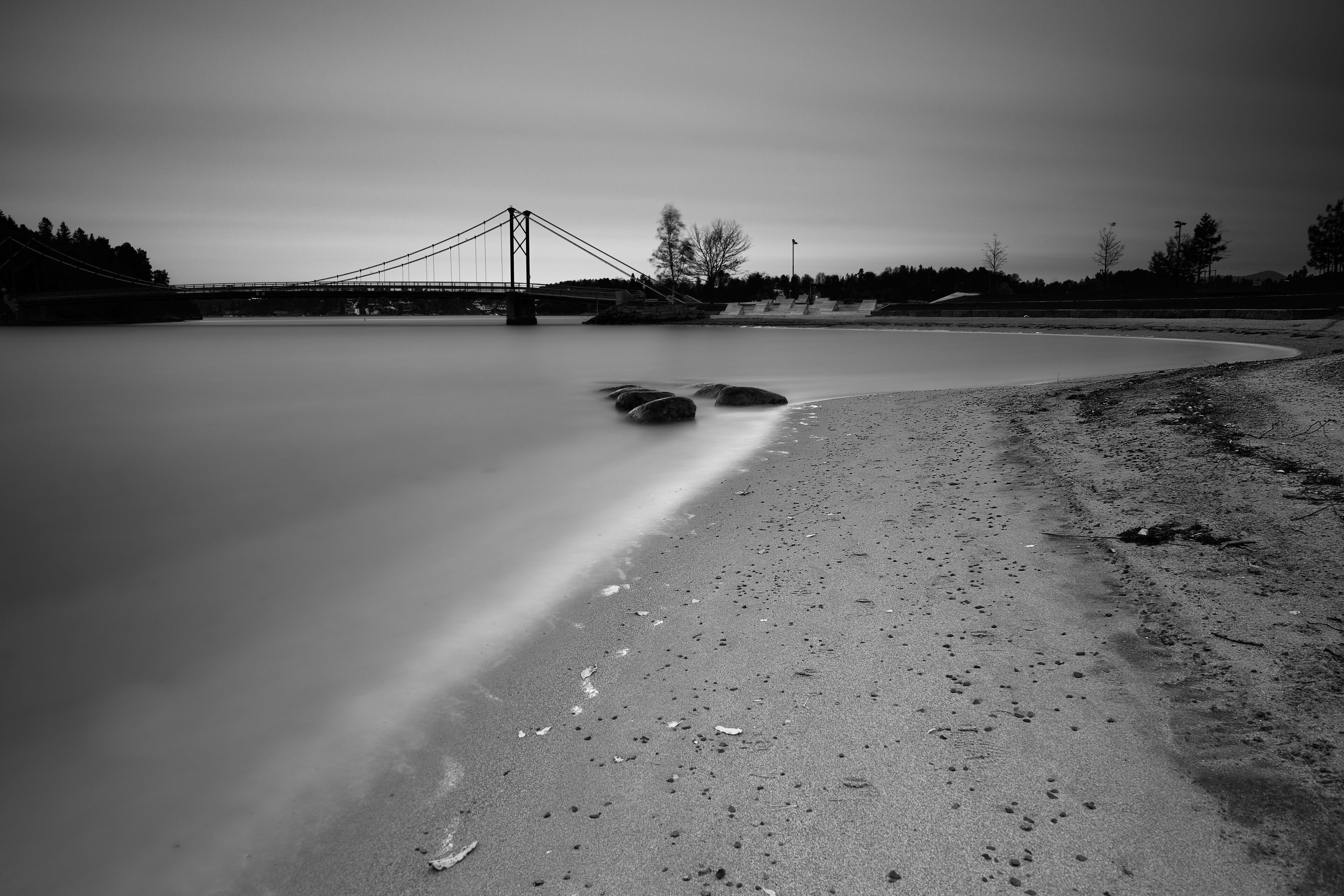 A bridge over a calm body of water with a sandy shore and rocks in the foreground.