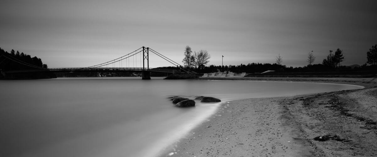 A bridge over a calm body of water with a sandy shore and rocks in the foreground.