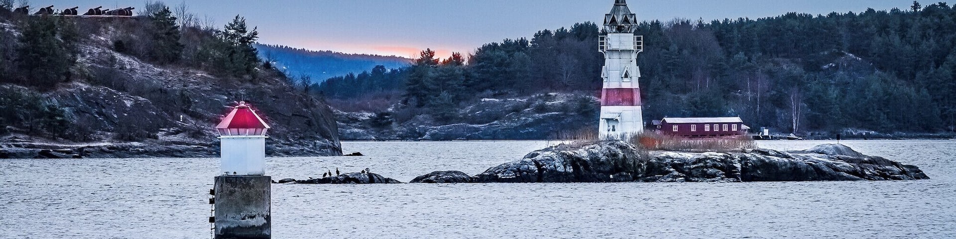 A lighthouse on a rocky outcrop with a red top, surrounded by snow-covered ground and a forested hill in the background.