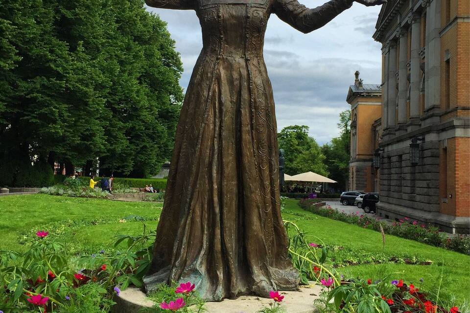 Une statue d’une femme vêtue d’une robe, les bras tendus, entourée de fleurs colorées et de verdure, avec un bâtiment en arrière-plan.