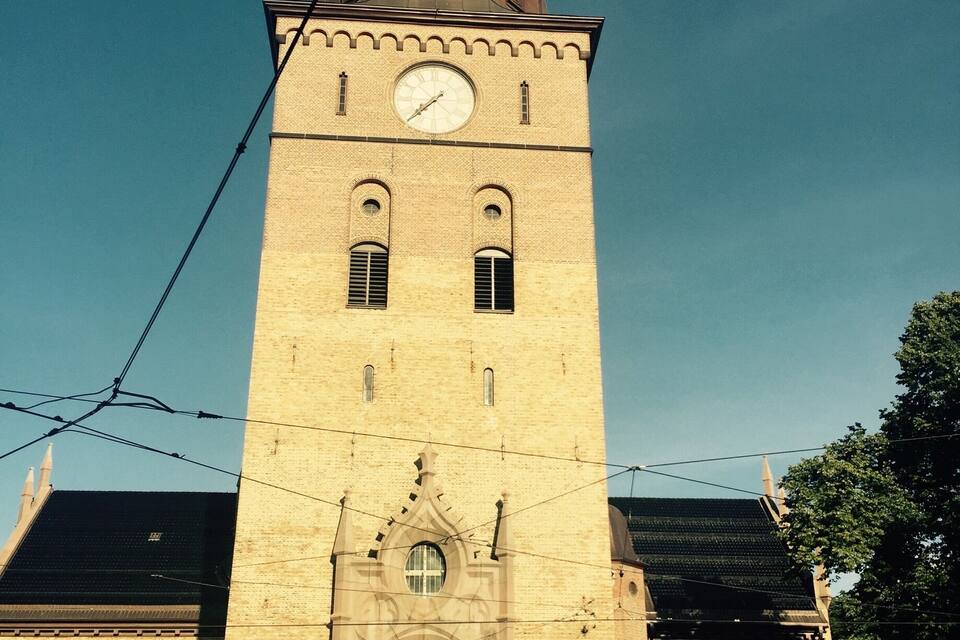 Une haute tour surmontée d’une horloge, dotée d’un toit en forme de flèche et d’une grande entrée en arc de cercle.