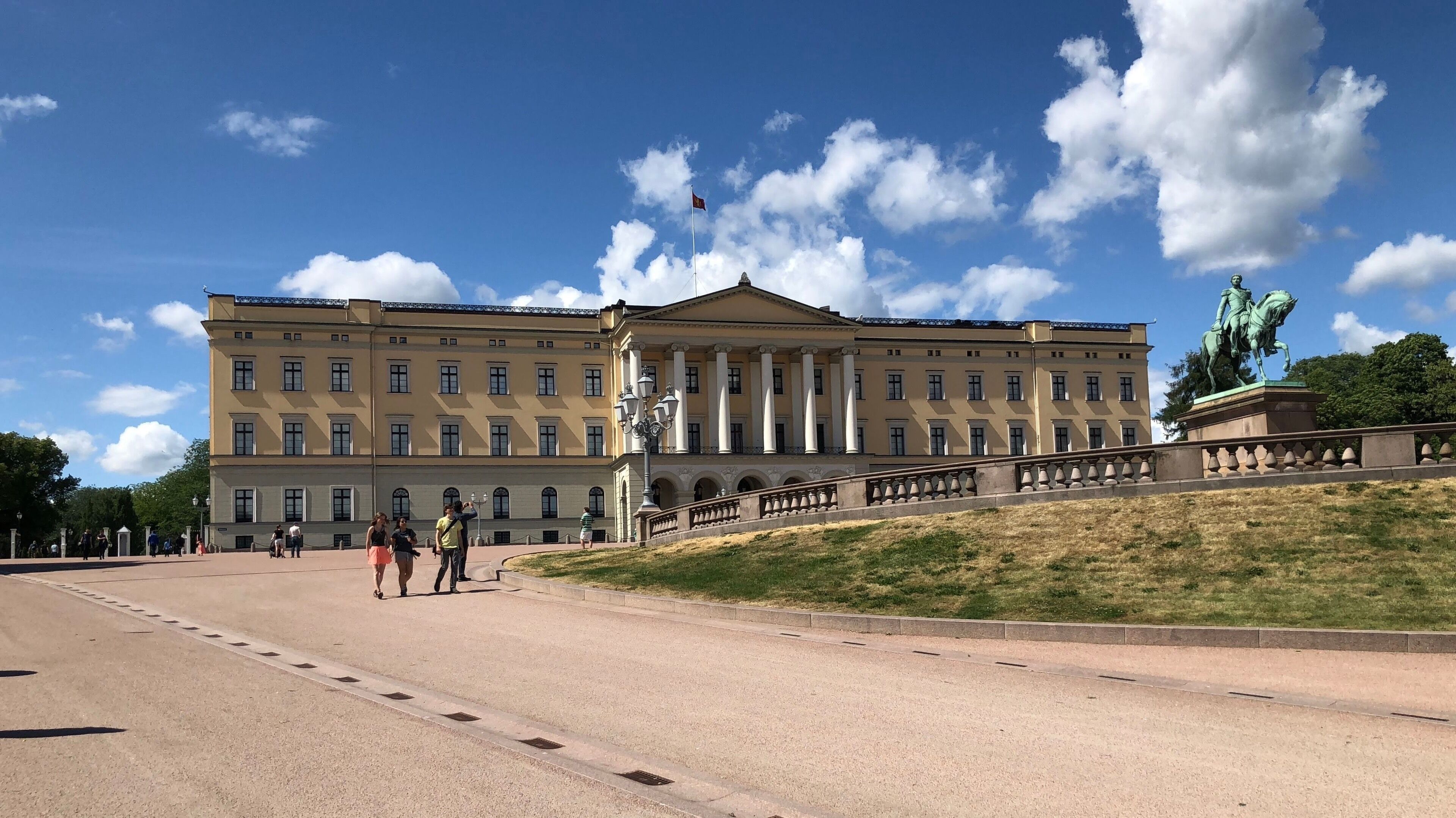 A large neoclassical building with a central pediment and columns, a statue of a figure on horseback, and a clear blue sky with scattered clouds.