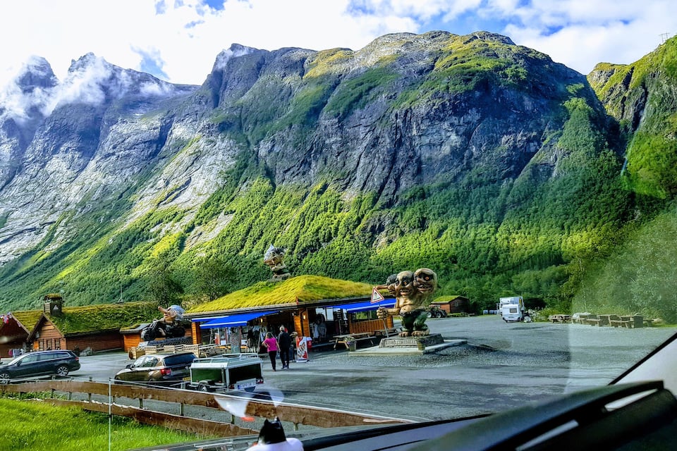A mountainous landscape with a parking area, a building with a blue roof, and a statue of a troll.