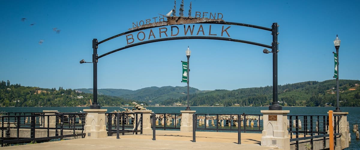 Segnaletica con la scritta 'North Bend Boardwalk' e vista su un lago e delle colline sullo sfondo.